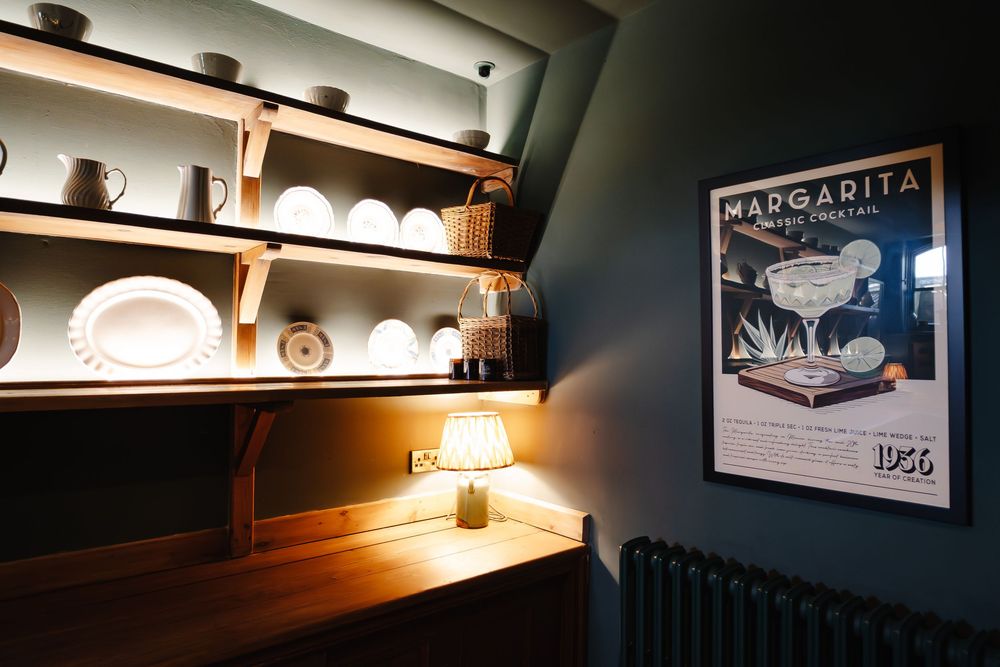 Cozy bar corner with wooden shelves displaying white dishes, warm lamp light, and a vintage Margarita cocktail poster on dark wall.
