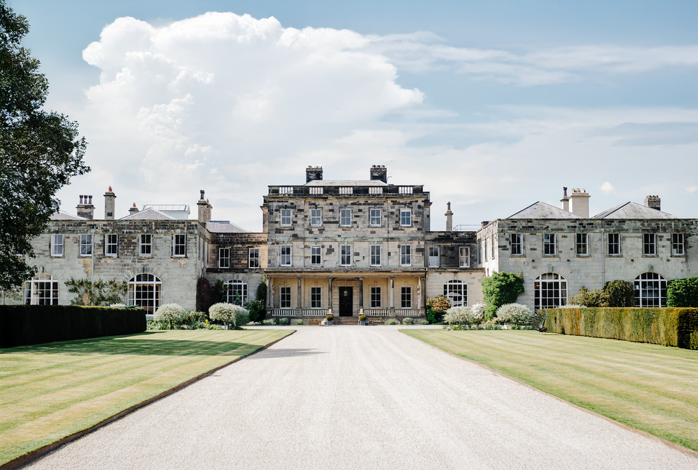 Grand English country manor house with sweeping lawn and gravel driveway under blue sky.