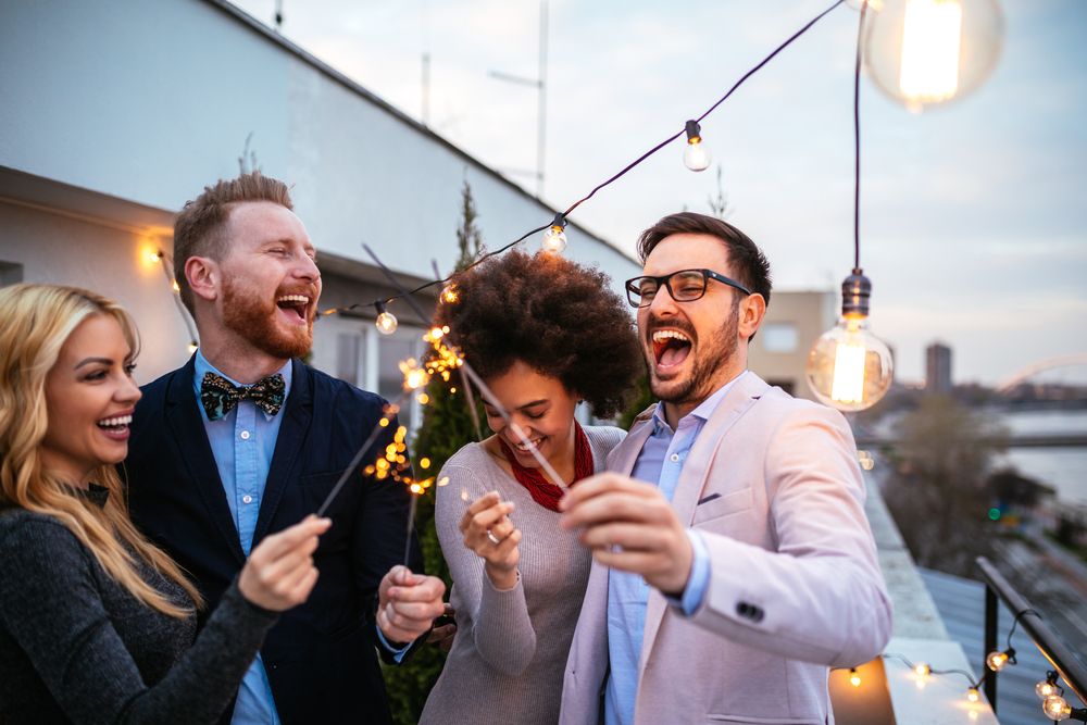 Friends celebrating with sparklers at a rooftop party, laughing and enjoying festive string lights at dusk.