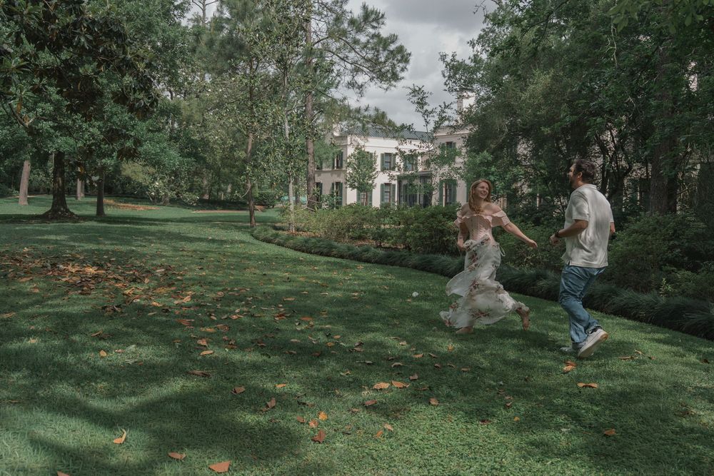 A playful couple runs hand-in-hand across a green lawn with a grand estate in the background.