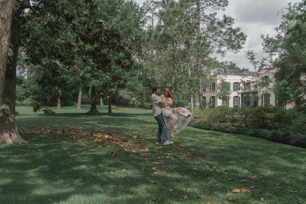 A man lifts his partner joyfully in a lush green garden beside a historic manor house.