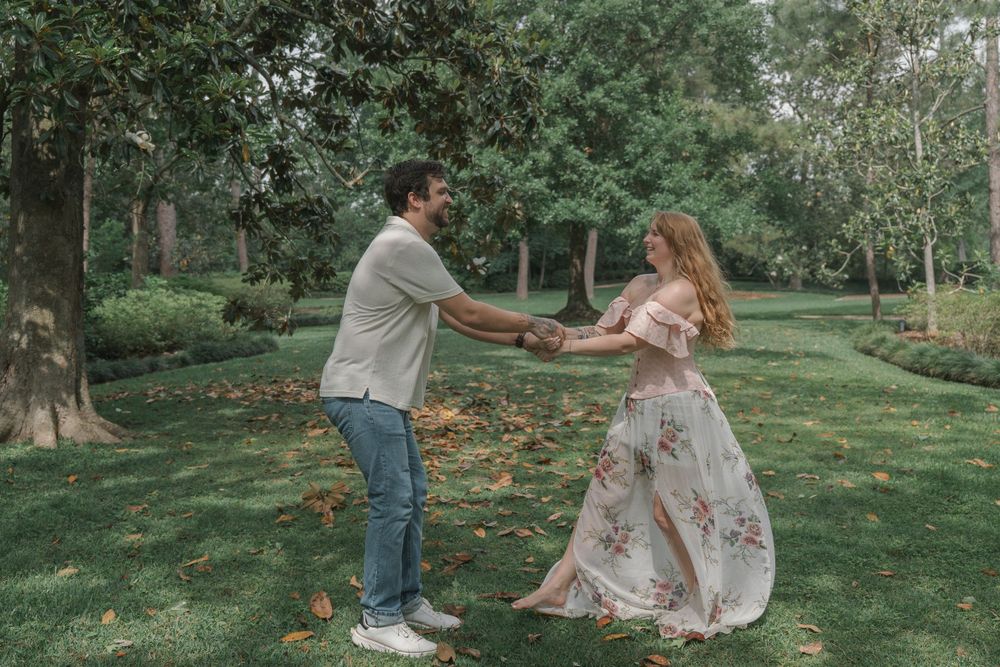 A couple holds hands and dances playfully in a scenic garden with autumn leaves on the ground.