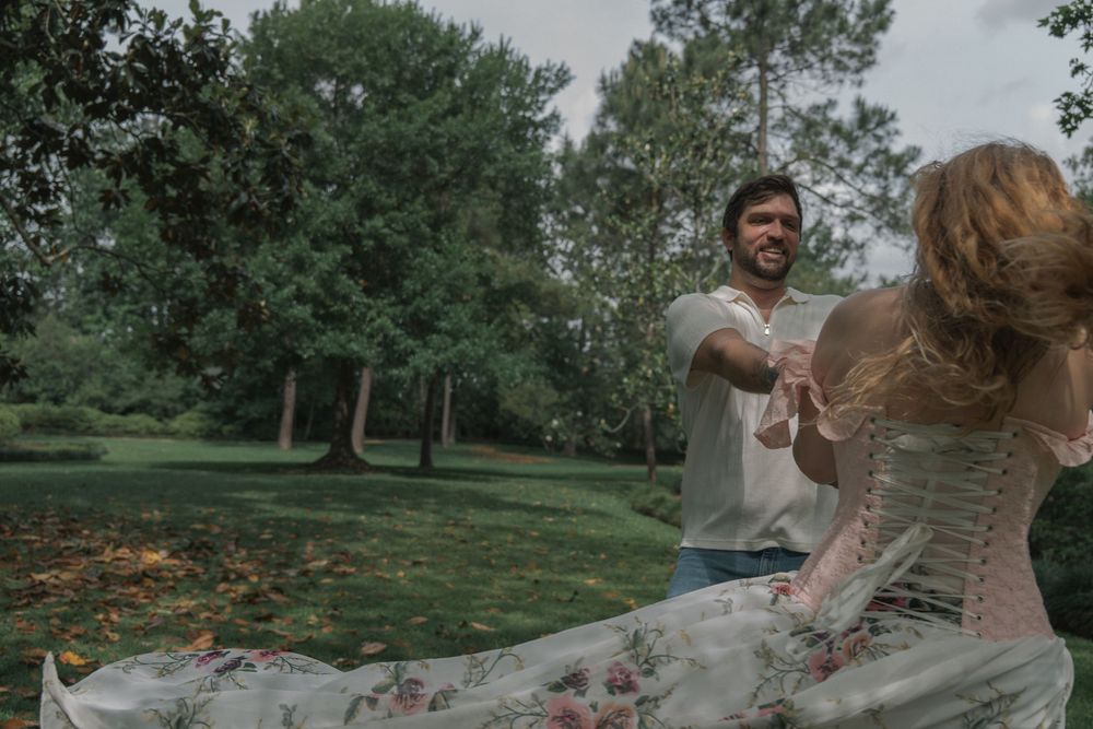 A bearded man and red-haired woman dance energetically together in a green park setting.