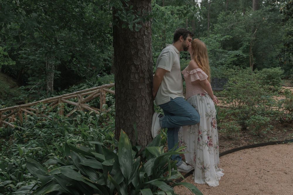 A romantic couple shares a kiss leaning against a tree on a scenic garden pathway surrounded by greenery.
