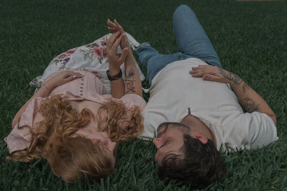 A couple lies head-to-head on lush green grass, sharing a quiet romantic moment outdoors.
