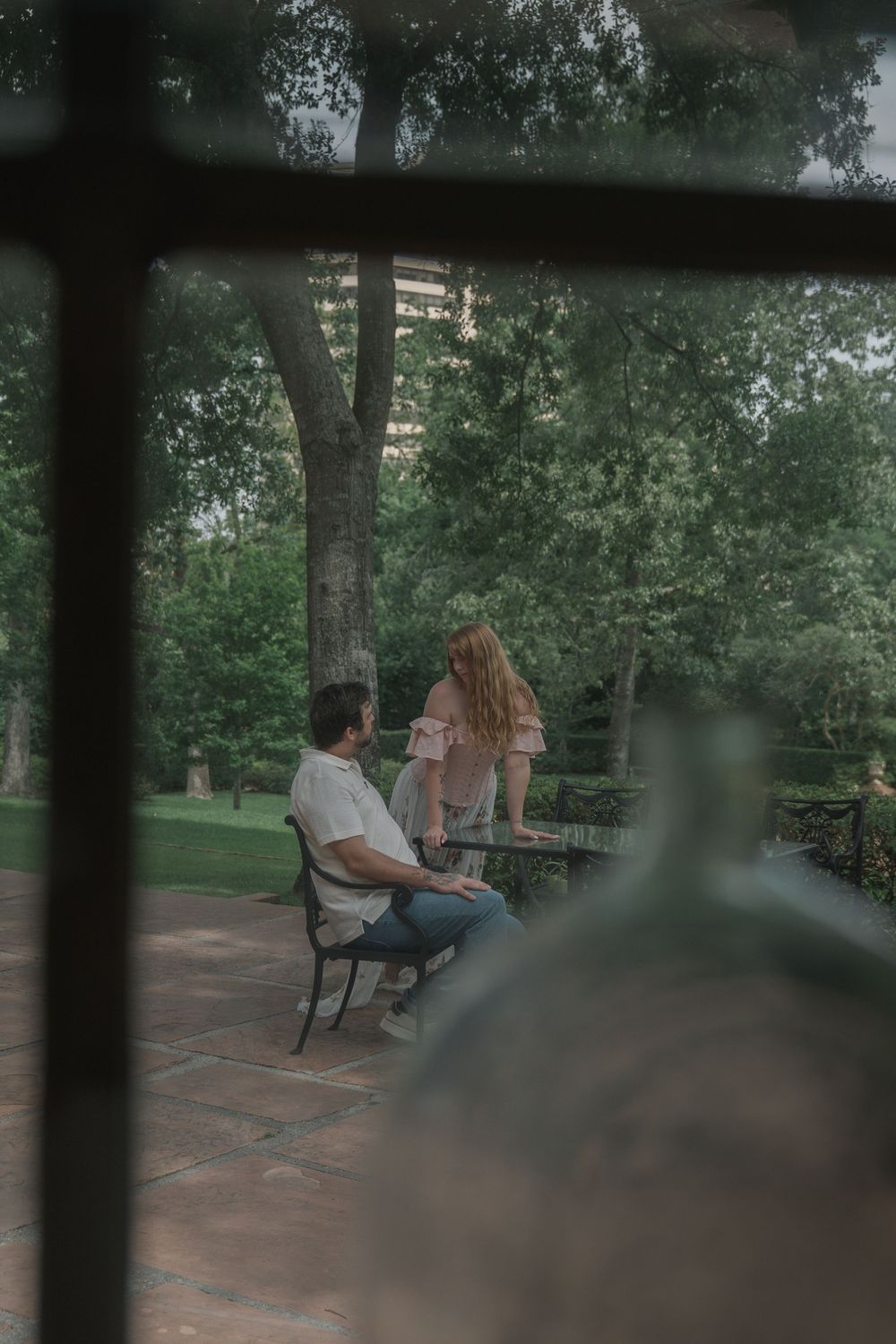 A couple enjoys a quiet moment in a lush garden, viewed through a window frame with bokeh.
