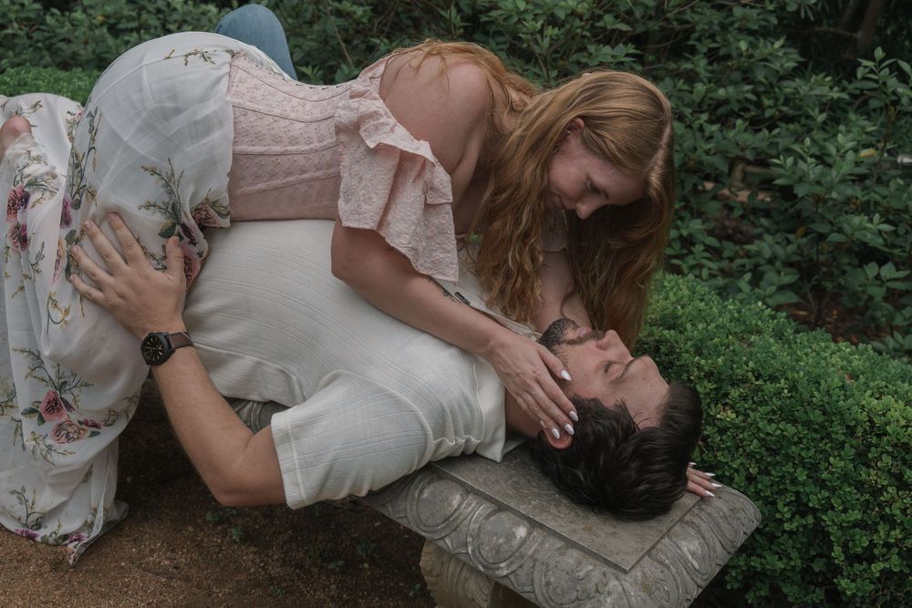A woman in a floral dress leans over a man lying on a stone bench in a garden setting.