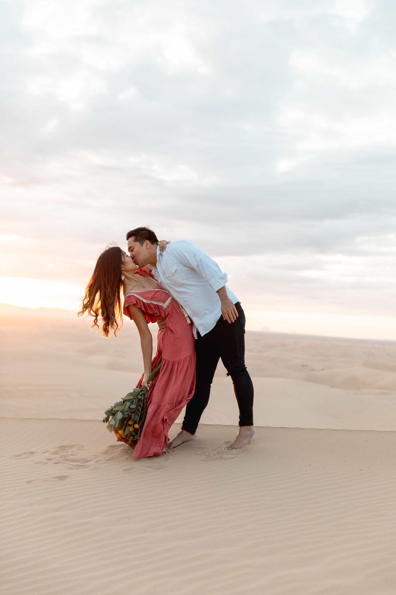 Couple sharing a romantic dip kiss on sandy dunes at desert sunset.