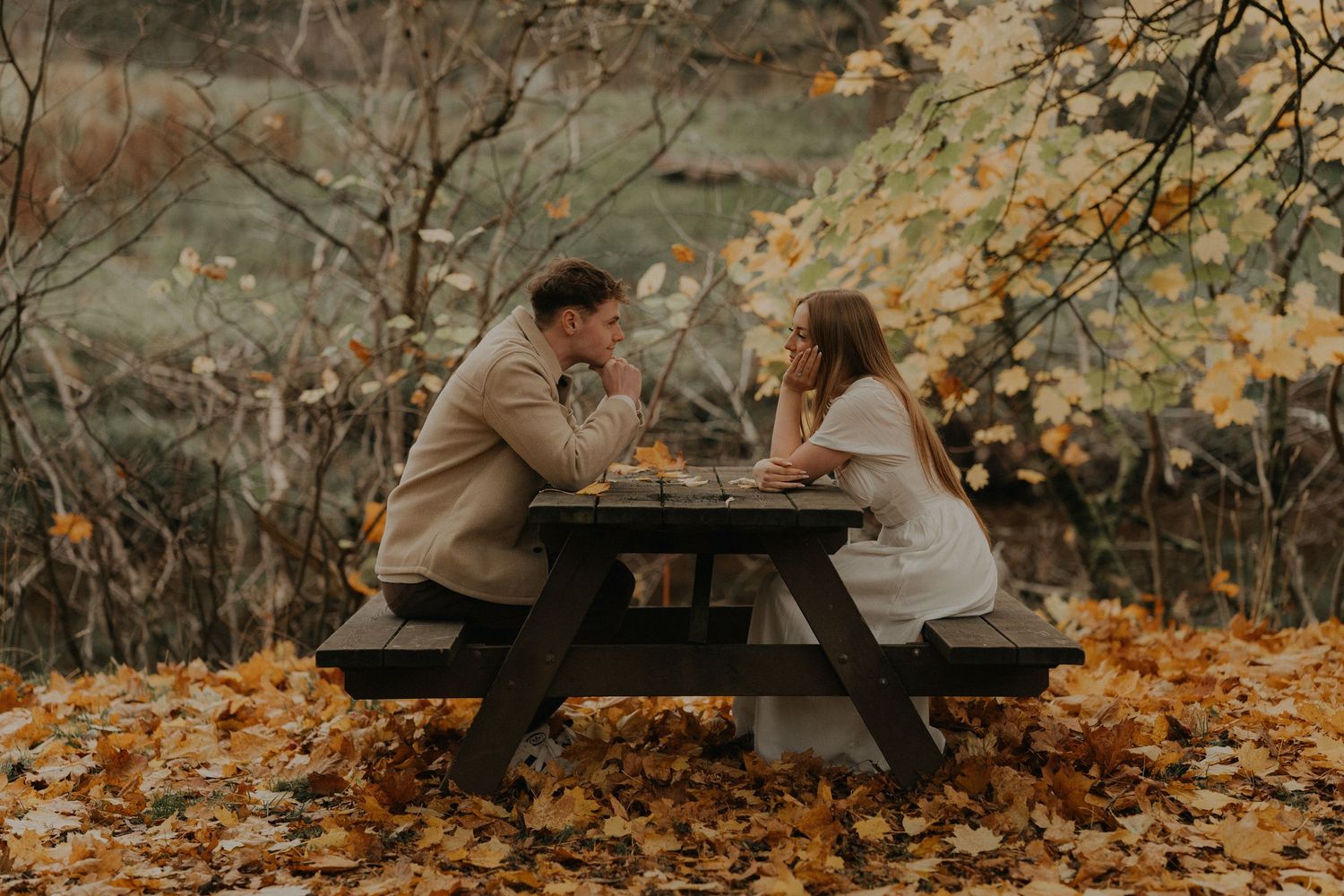 Couple sitting across from each other at a picnic table surrounded by golden autumn leaves in a romantic outdoor setting.