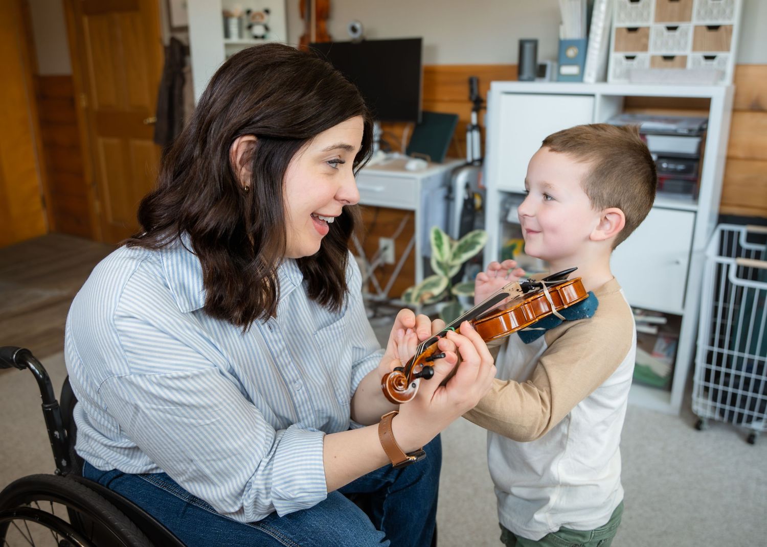 Woman in wheelchair smiling with young boy playing violin together in a bright home setting.