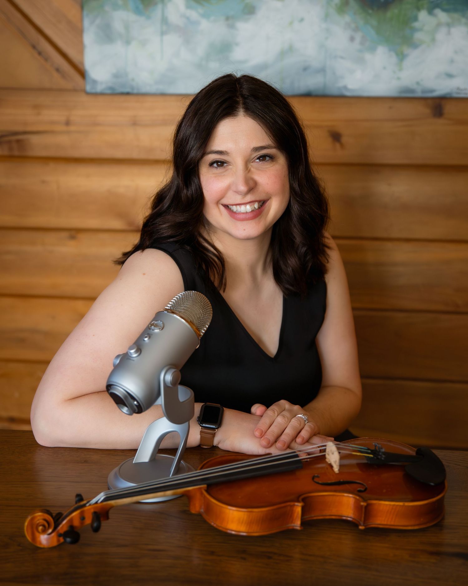 Smiling woman with dark hair poses with a violin and microphone on a wooden table in a cozy studio setting.
