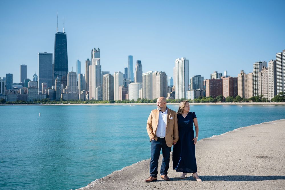 Couple walking along Chicago lakefront with city skyline and John Hancock Center in background on sunny day.