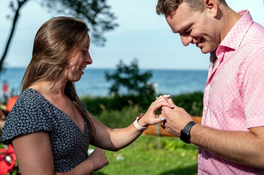 Man in pink shirt placing ring on womans finger outdoors near lake during romantic marriage proposal.