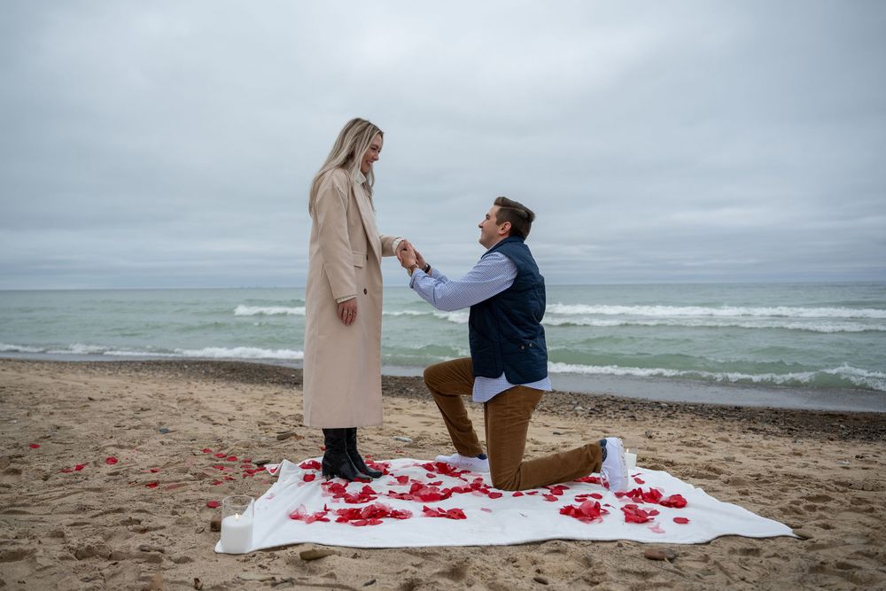 Man proposes on a rose petal-covered beach blanket to a woman on a cloudy day by the ocean.