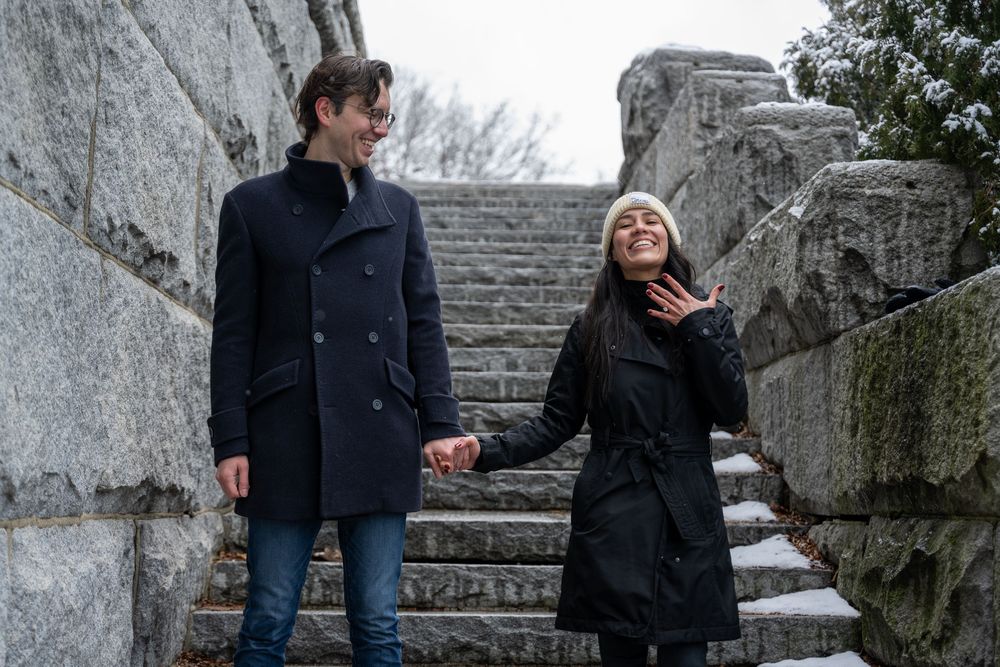 Couple holding hands and smiling on stone steps outdoors in winter, wearing dark coats in a romantic engagement photo.