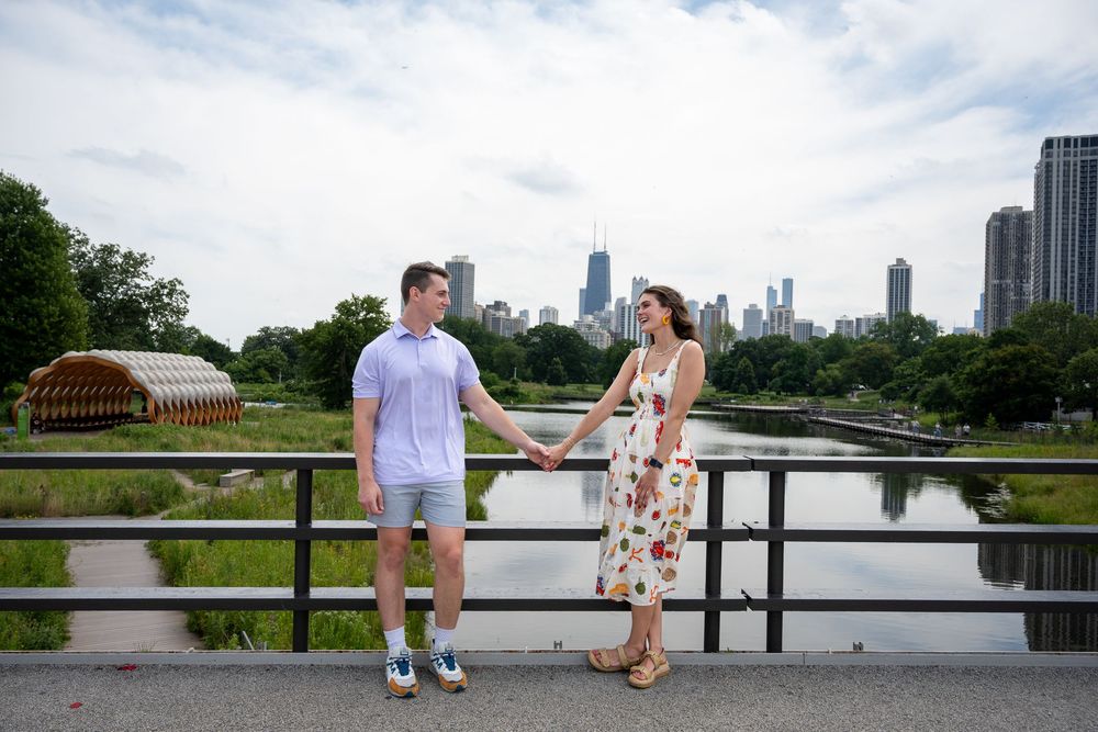 Couple holds hands on a bridge at Lincoln Park Zoo with Chicago skyline in the background on a cloudy day.