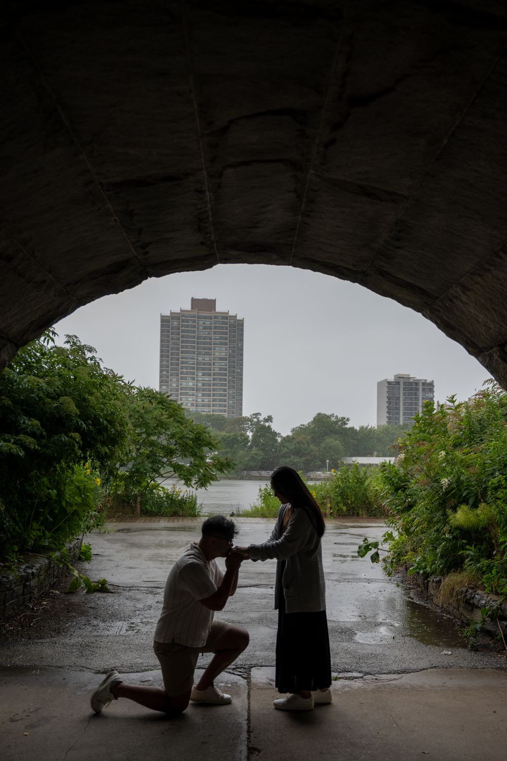 Man proposes on one knee under stone arch bridge with city skyline in background on a rainy day.