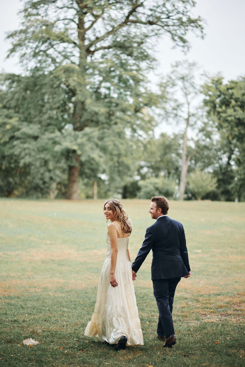 Bride and groom walking hand in hand through a lush green park, bride in flowing gown glancing back smiling.