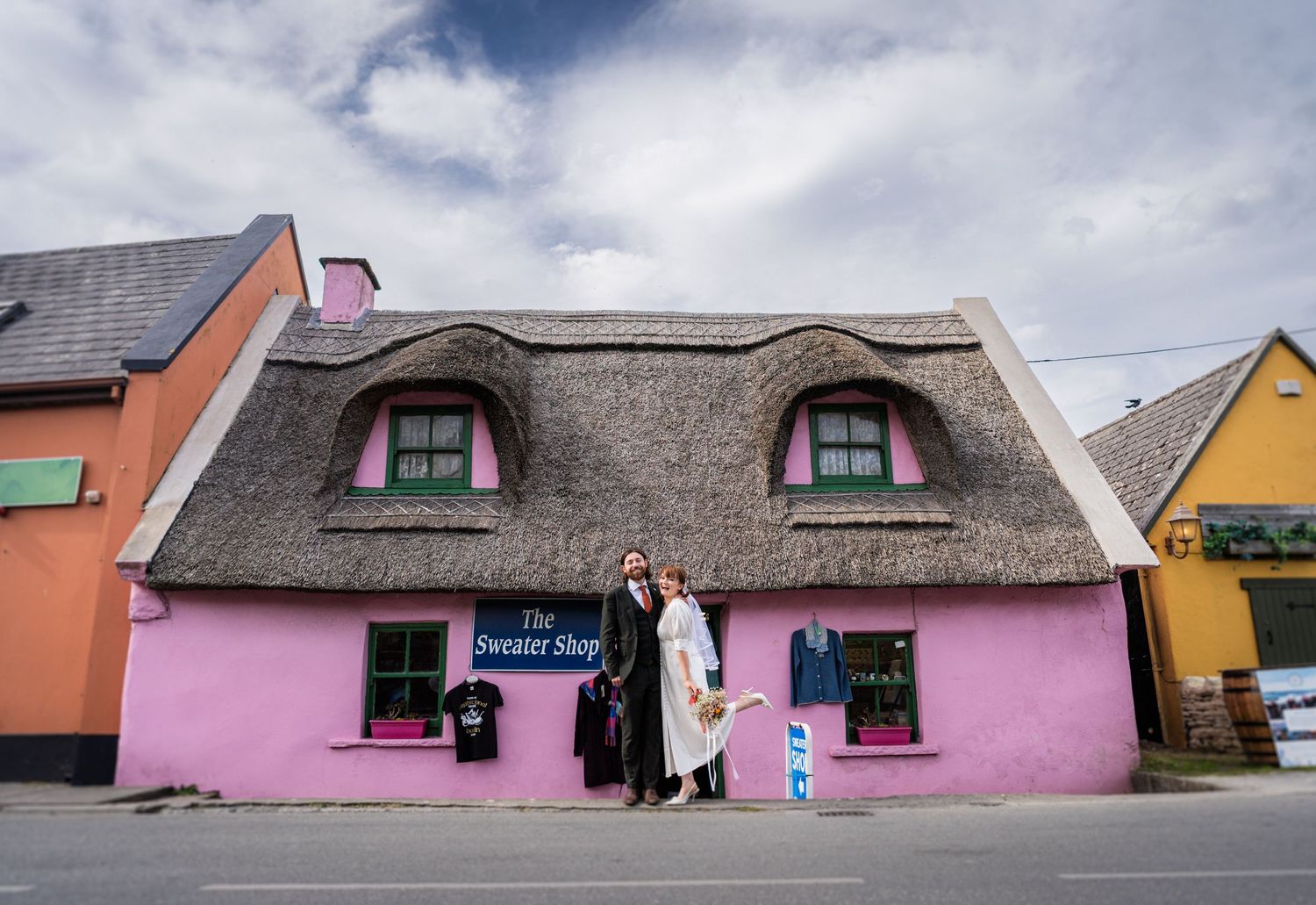 Two people stand outside The Sweater Shop, a pink thatched-roof cottage in a charming Irish village street.