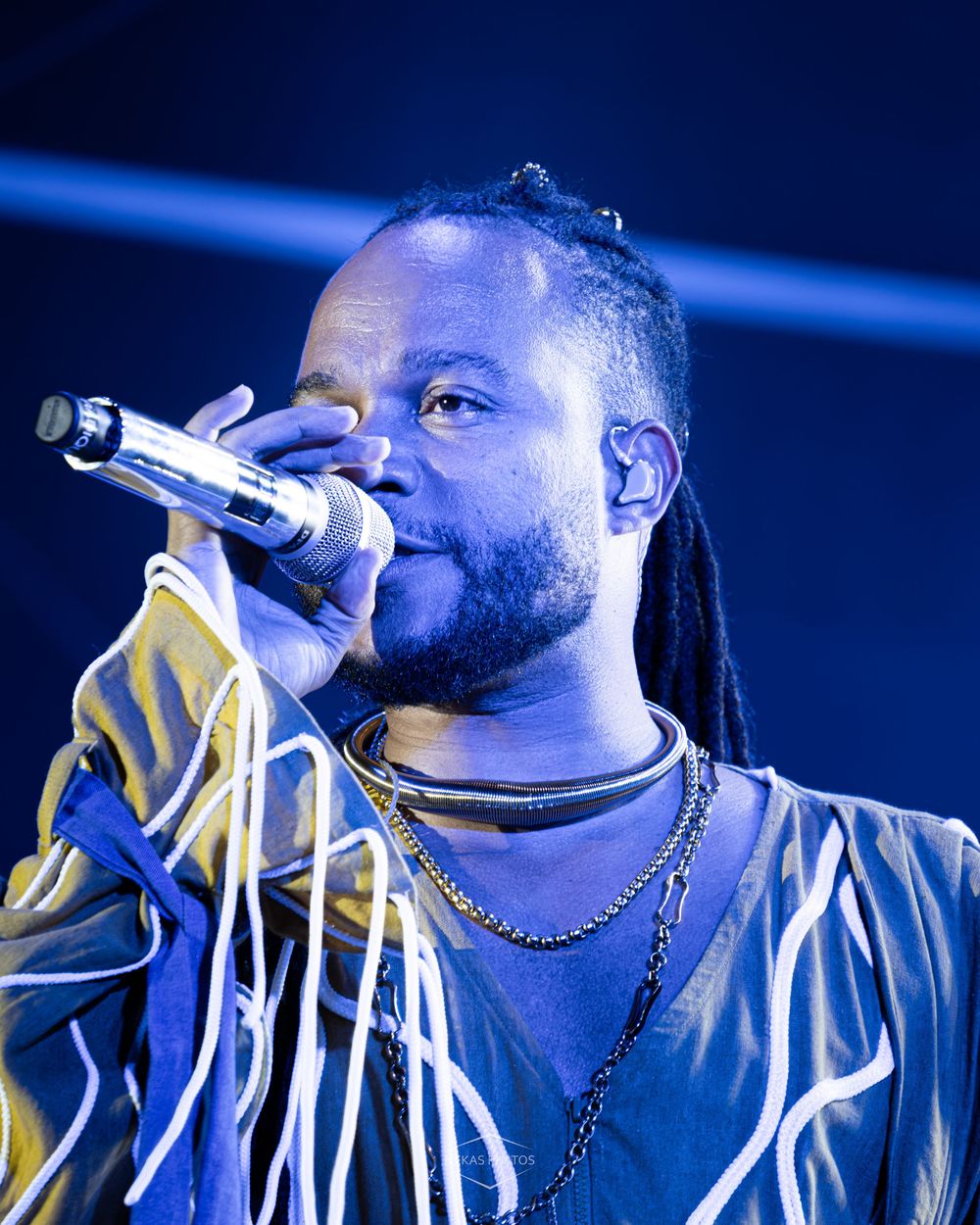 A male performer with dreadlocks sings into a microphone on stage, bathed in blue light, wearing a striped jacket.