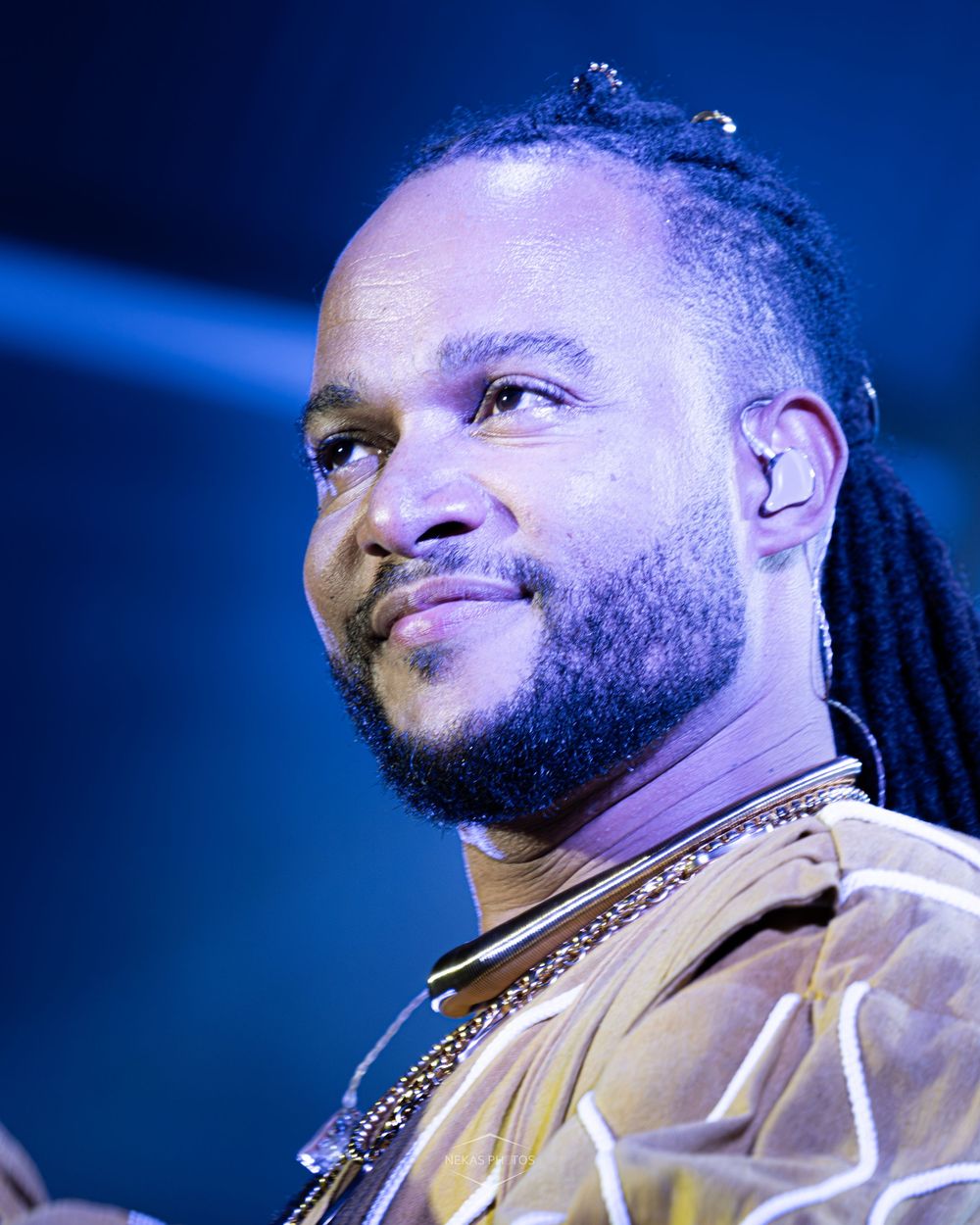 A man with dreadlocks and a beard performs on stage, wearing a gold jacket and chain necklace under blue lights.