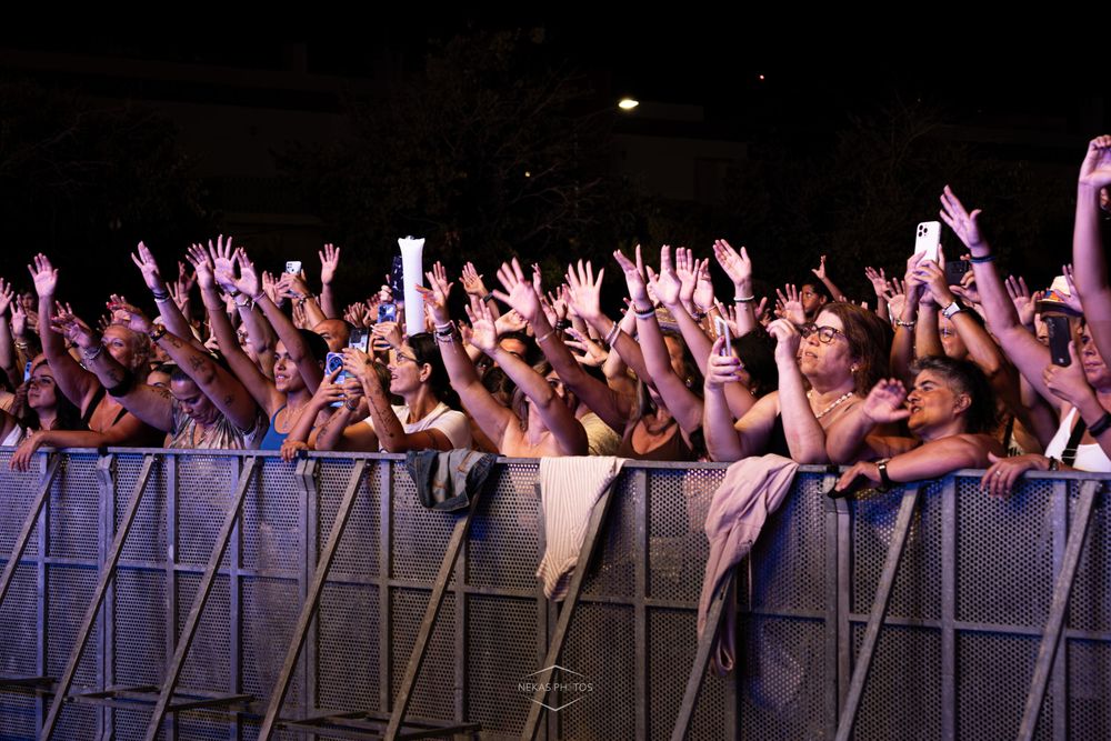 Large crowd of fans raising hands and phones at outdoor concert, pressing against security barrier at night.