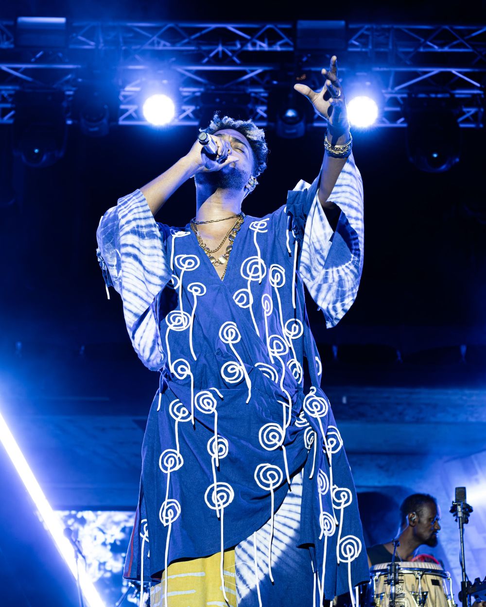 Performer in blue patterned African kaftan singing on stage with dramatic lighting at an outdoor concert.
