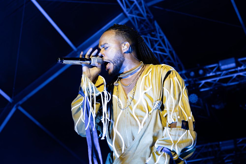 Male singer performing on stage wearing gold satin outfit, holding microphone with dramatic lighting at concert.