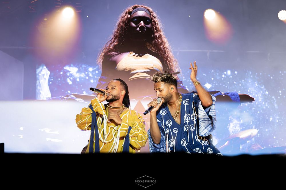 Two male performers in colorful African attire sing on stage with a large projected image behind them.