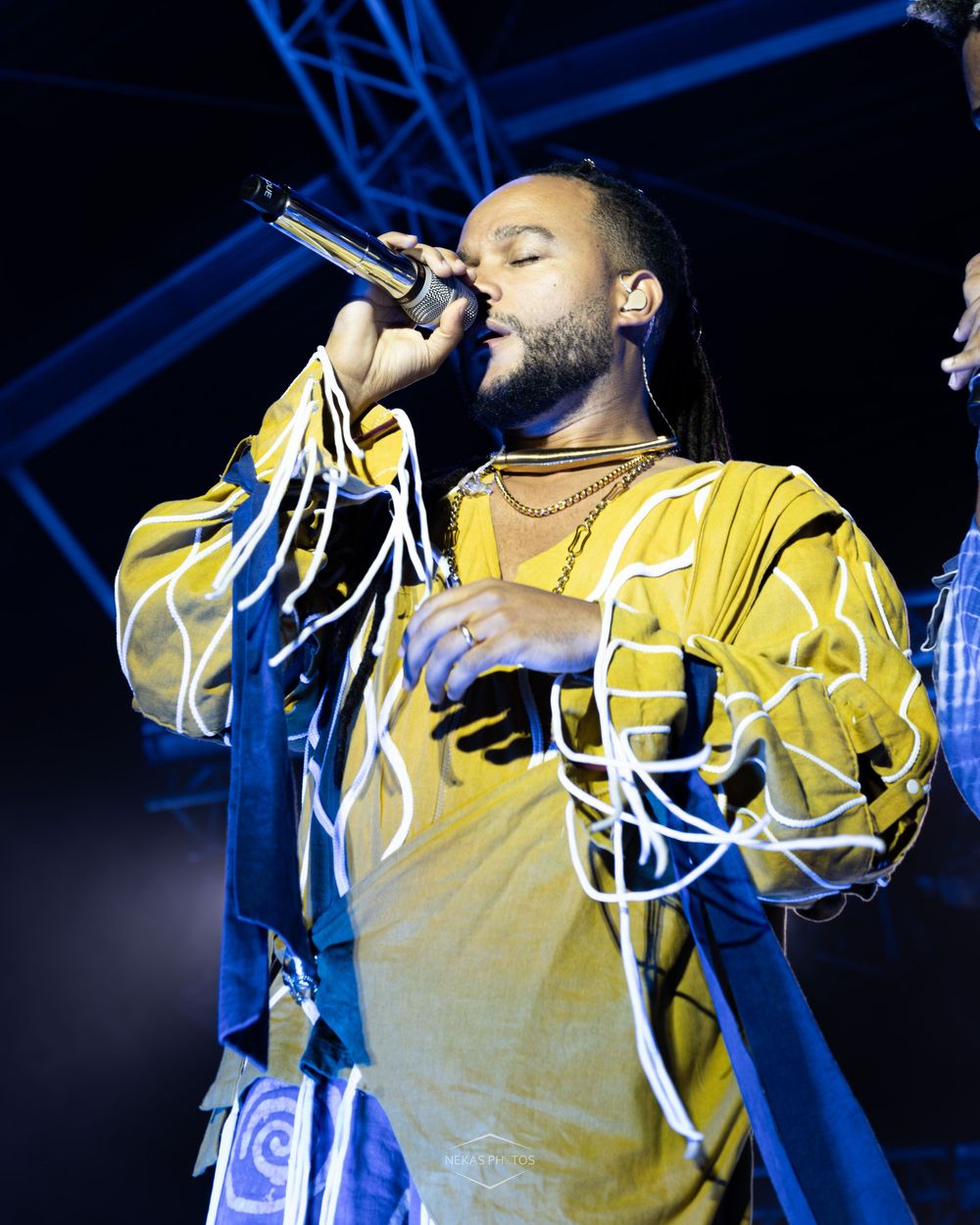 A male performer with dreadlocks sings into a microphone on stage wearing a yellow outfit with white rope details.