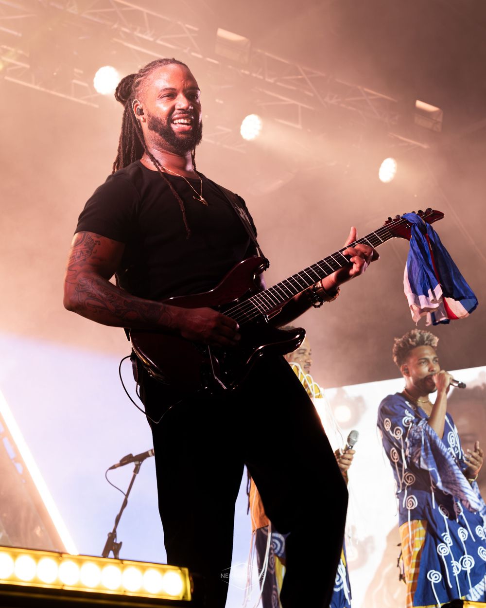 Musician with dreadlocks plays electric guitar on stage under bright lights during a live concert performance.