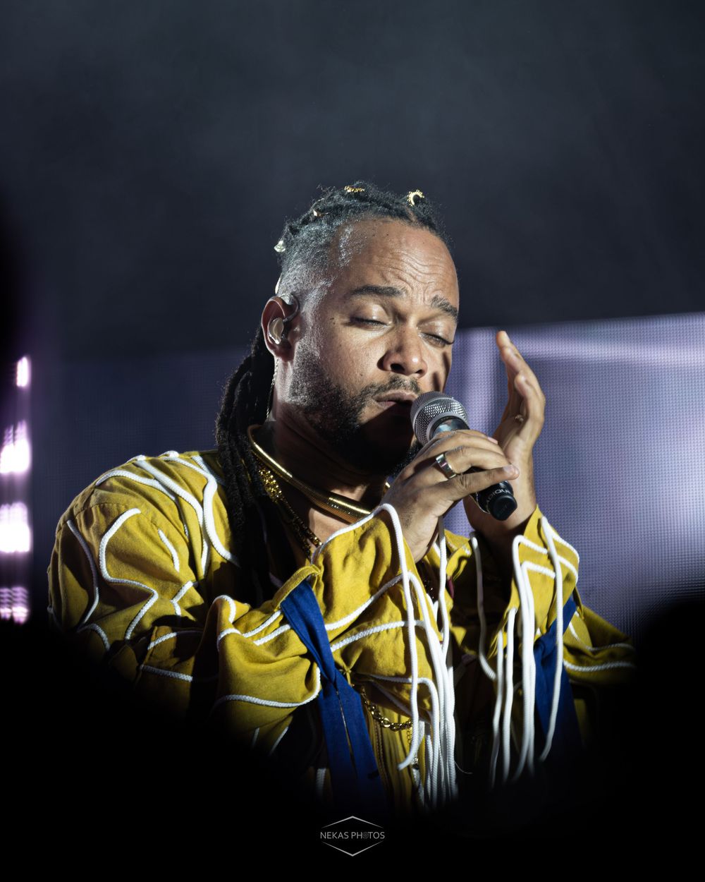 Male singer with dreadlocks performing on stage wearing a yellow patterned shirt, holding a microphone with eyes closed.