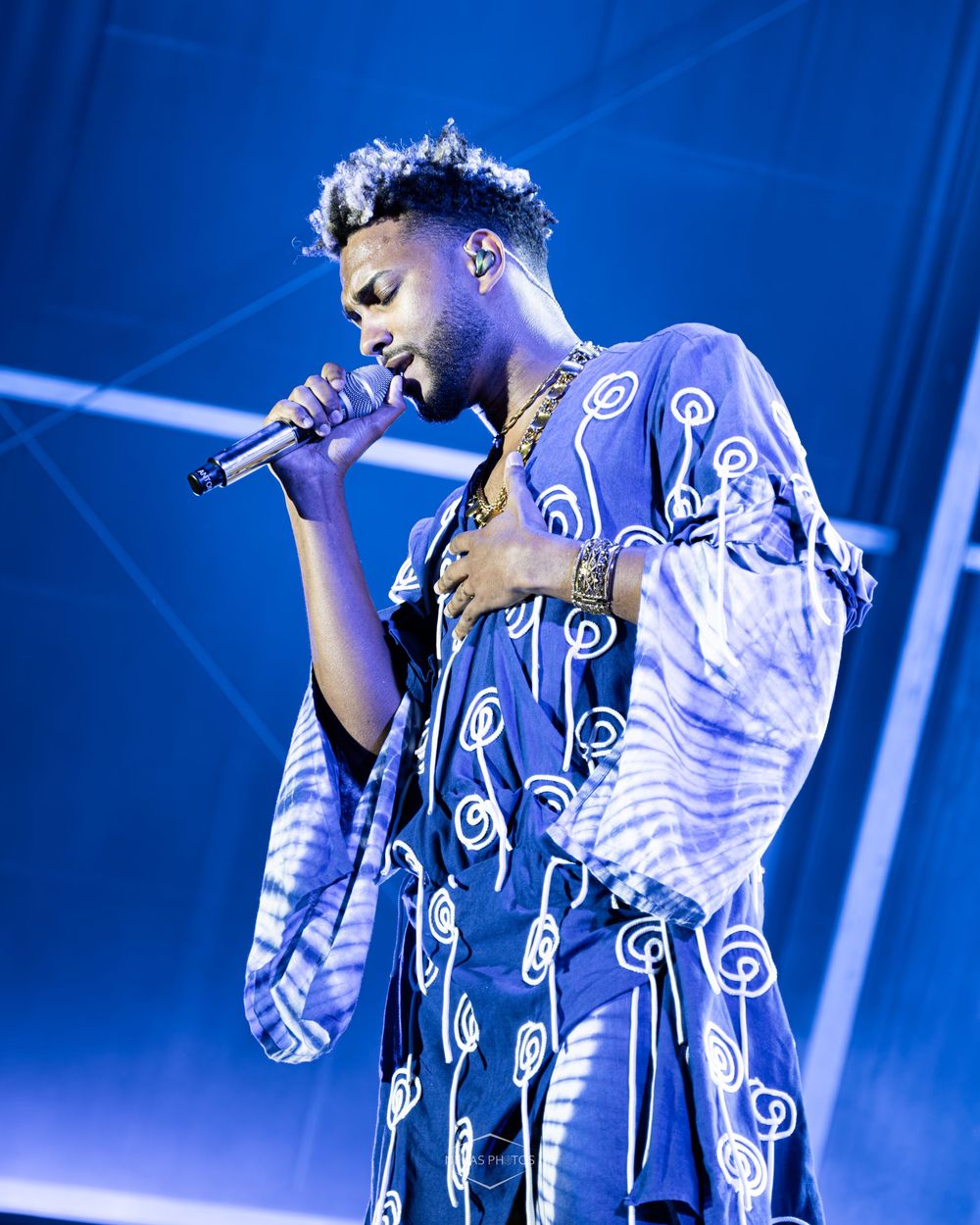 A young male singer performs on stage wearing a blue patterned outfit, holding a microphone under blue stage lighting.