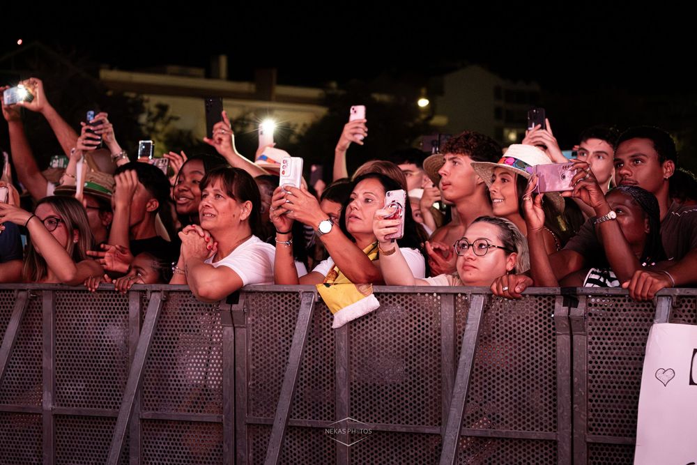 Excited crowd of fans holding smartphones at an outdoor concert at night, pressed against a metal barrier.