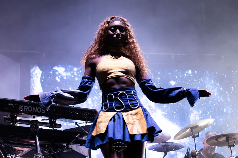 Performer in blue costume with long hair on stage under dramatic lighting with keyboard and drums.