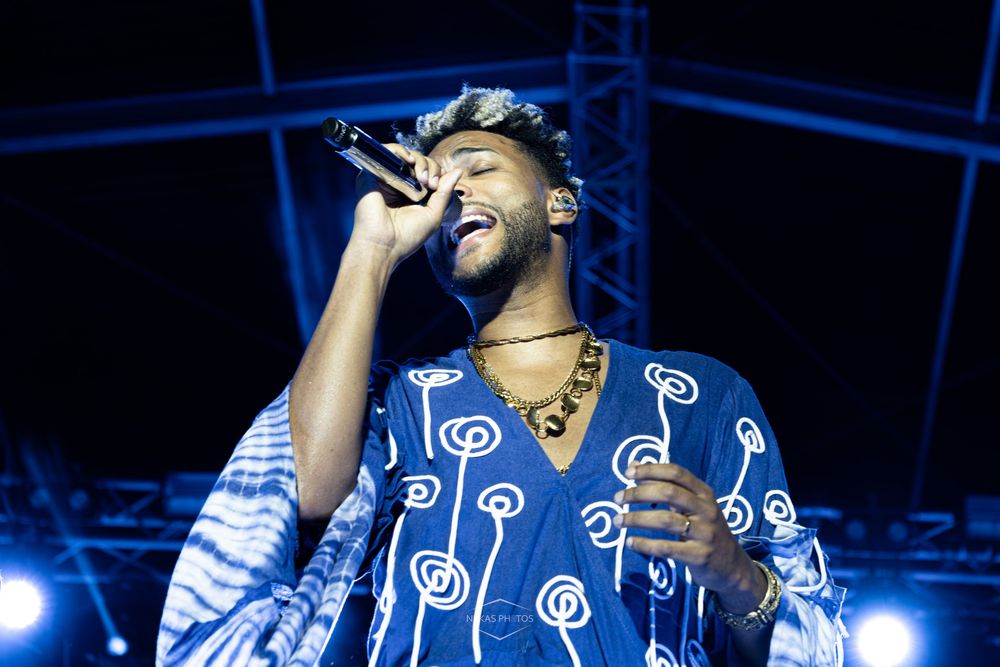 A male singer performs on stage wearing a blue patterned outfit, singing passionately into a microphone under stage lights.