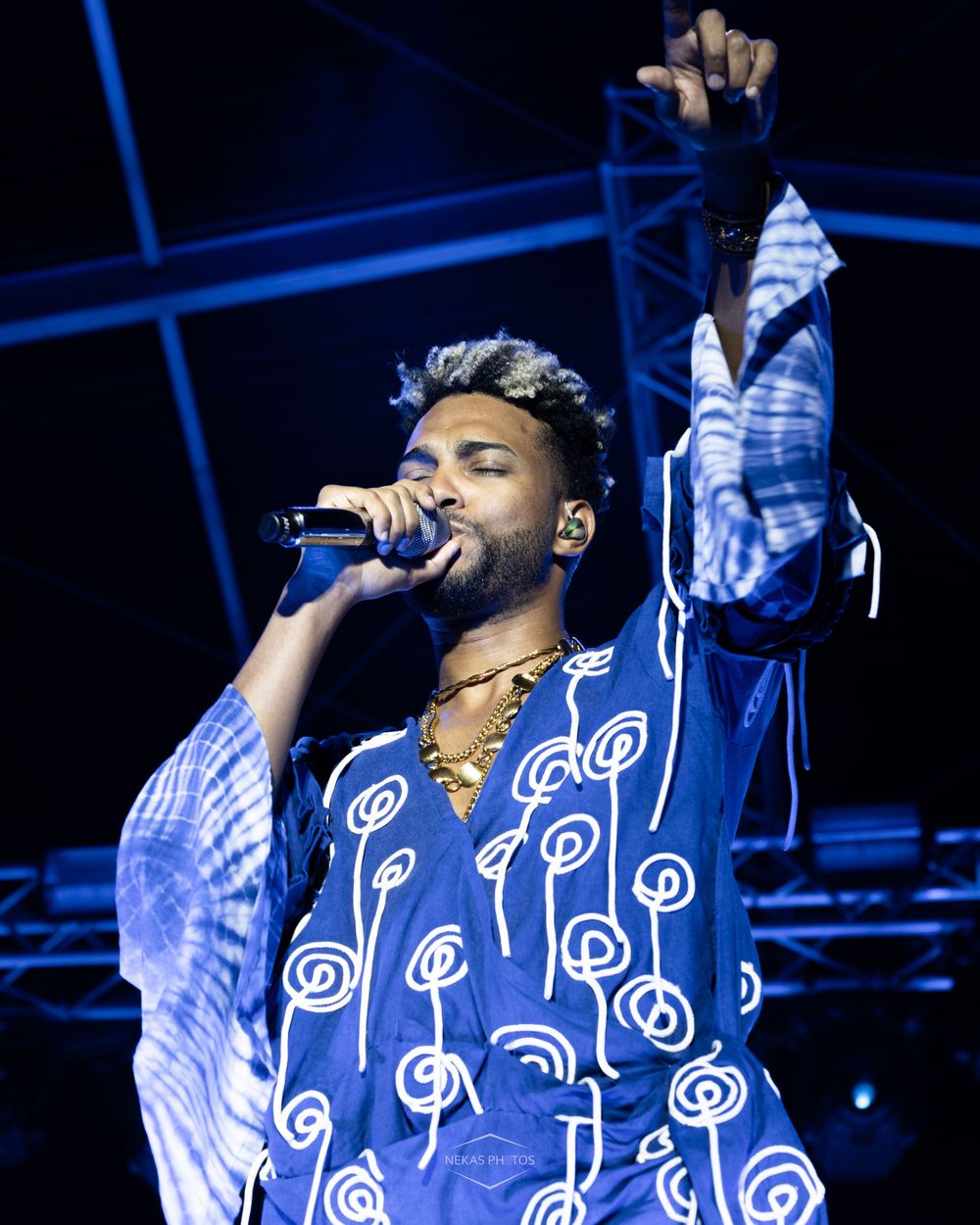 A male singer performs on stage holding a microphone, wearing a blue patterned robe with one hand raised under blue stage lighting.