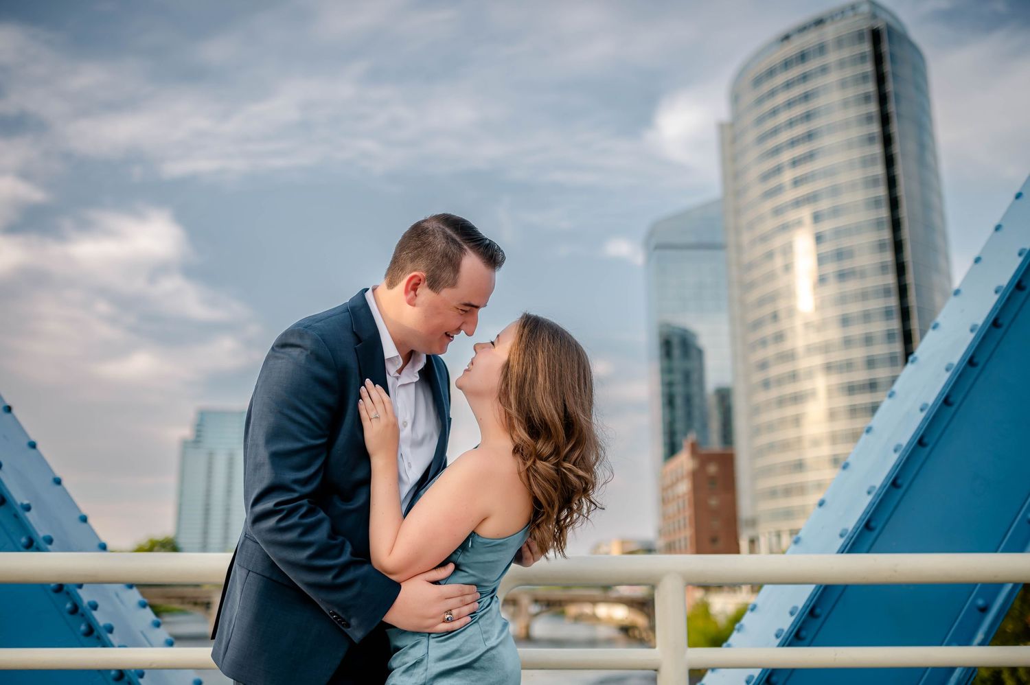 Couple sharing a romantic kiss on a blue bridge with Grand Rapids city skyline in the background.