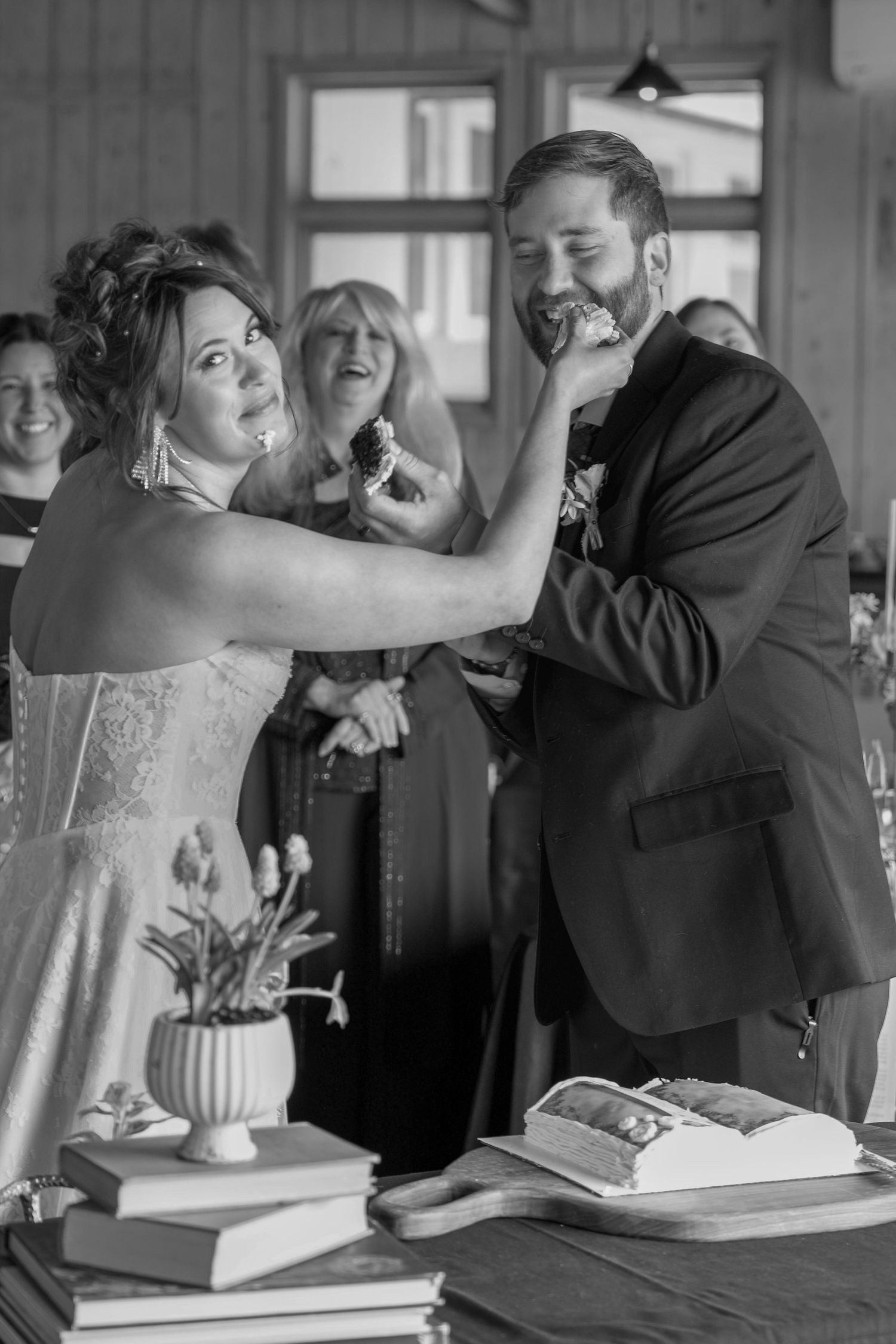 Black and white photo of bride feeding groom wedding cake at reception while guests laugh in background.