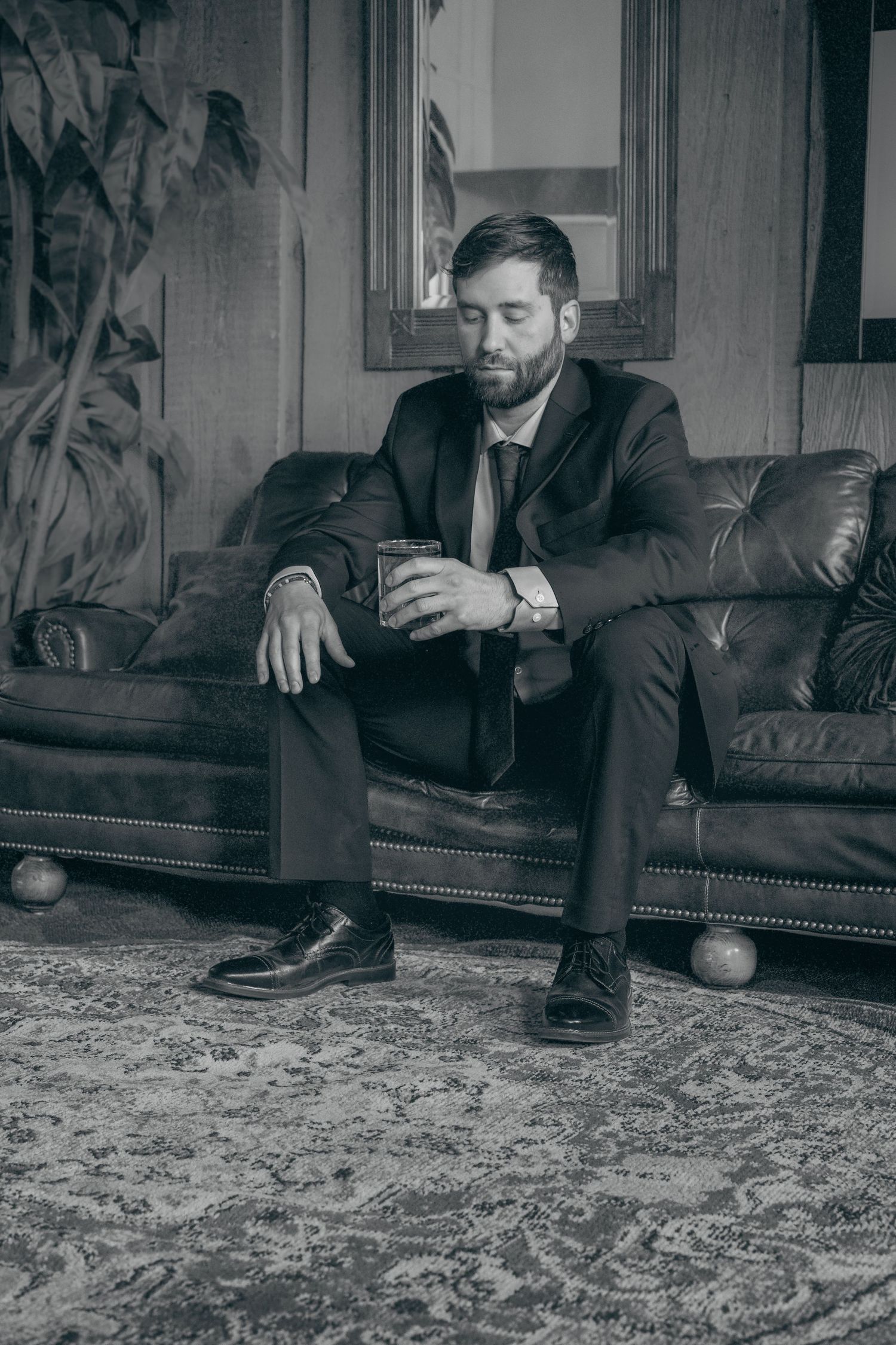 Black and white photo of a bearded man in a suit sitting on a leather sofa holding a glass.