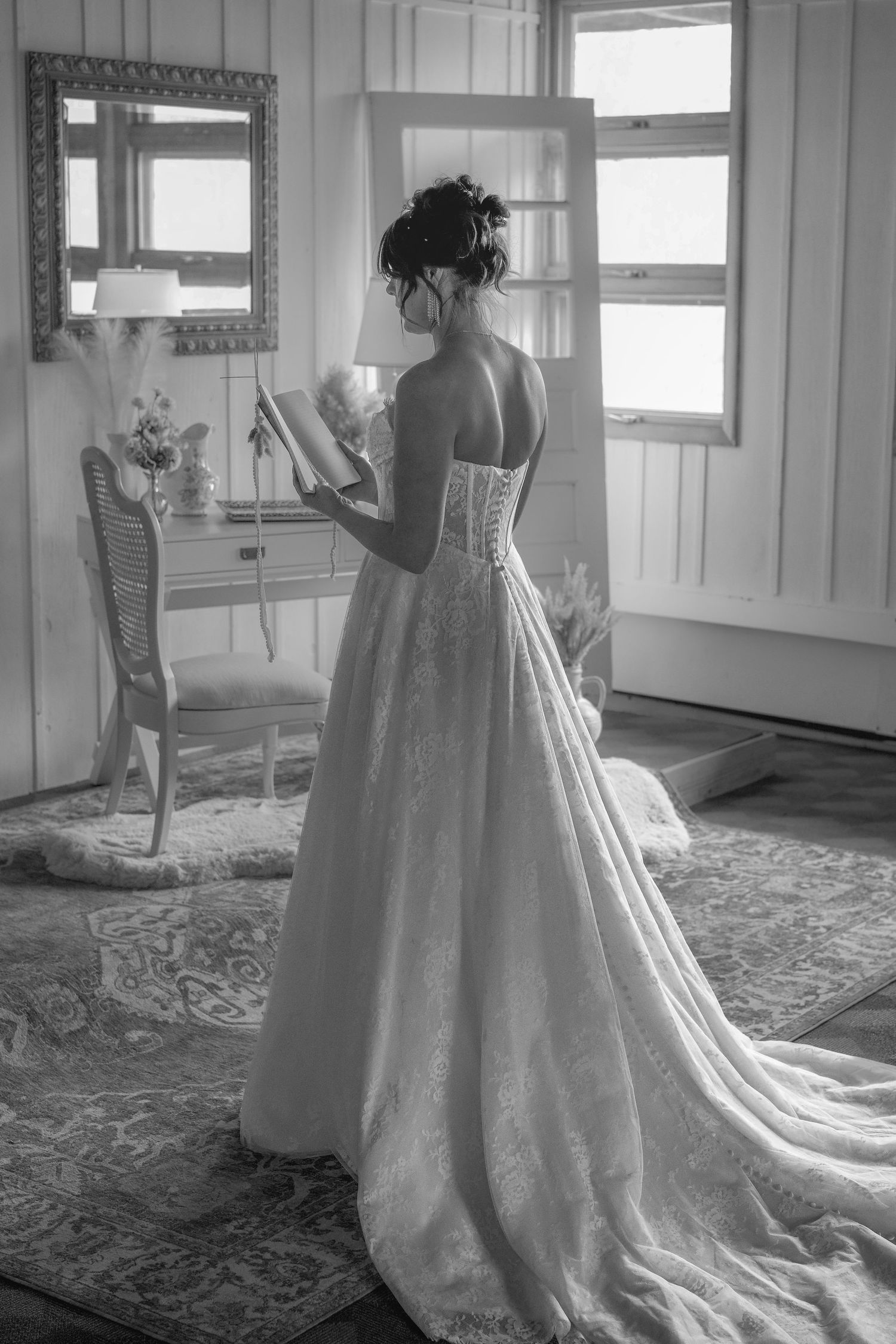 Black and white photo of a bride in a lace wedding gown with corset back, holding her veil in an elegant room.