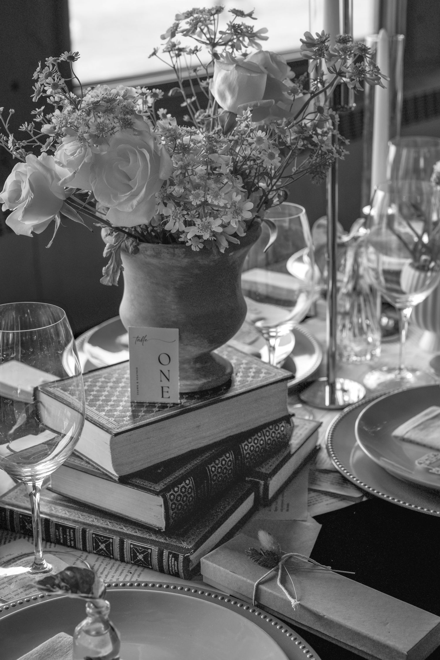 Black and white photo of elegant table setting with stacked books, floral centerpiece in urn vase, and crystal wine glasses.