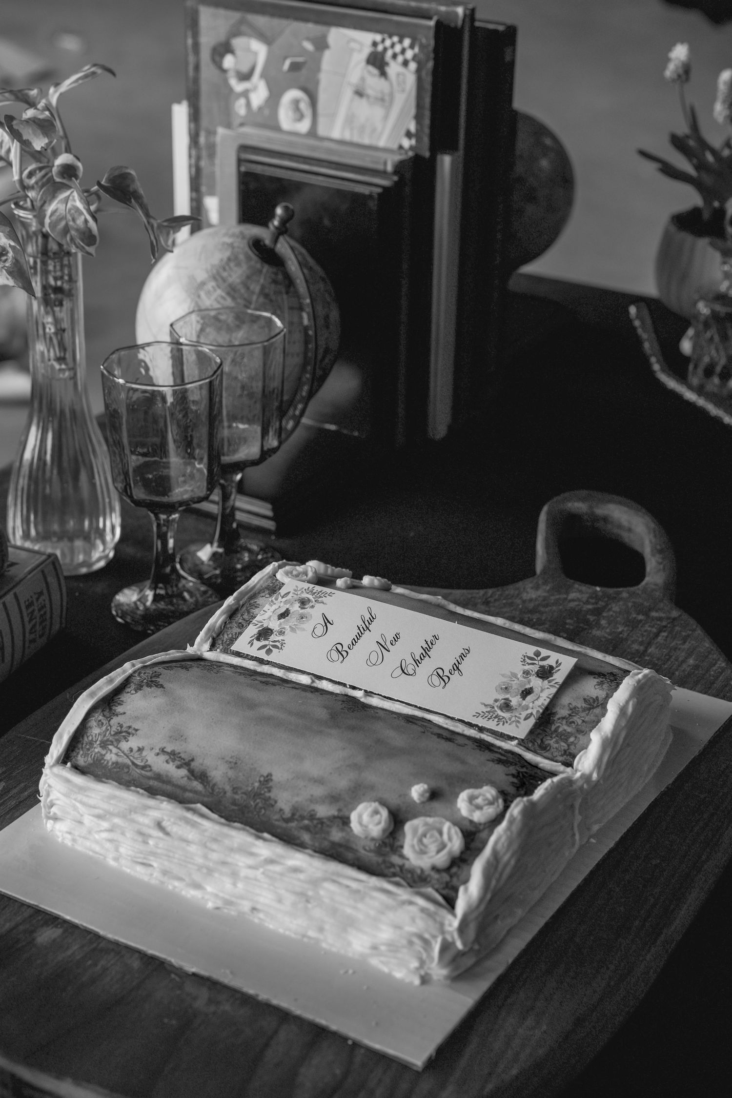 Black and white photo of a book-shaped cake with floral decor, globe, and vintage items on a wooden table.