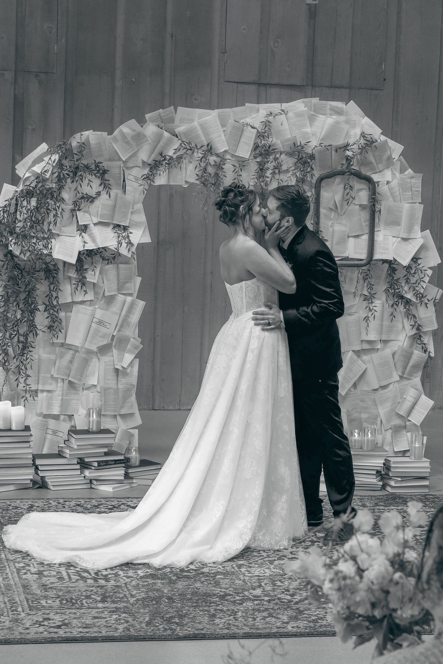 Black and white photo of bride and groom sharing first kiss under decorative arch made of book pages at wedding ceremony.