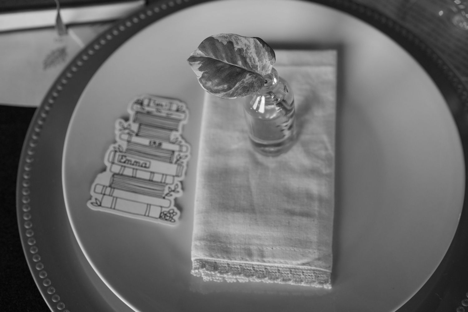 Black and white photo of an elegant place setting with a folded napkin, small vase, and decorative book stack cutout on a charger plate.