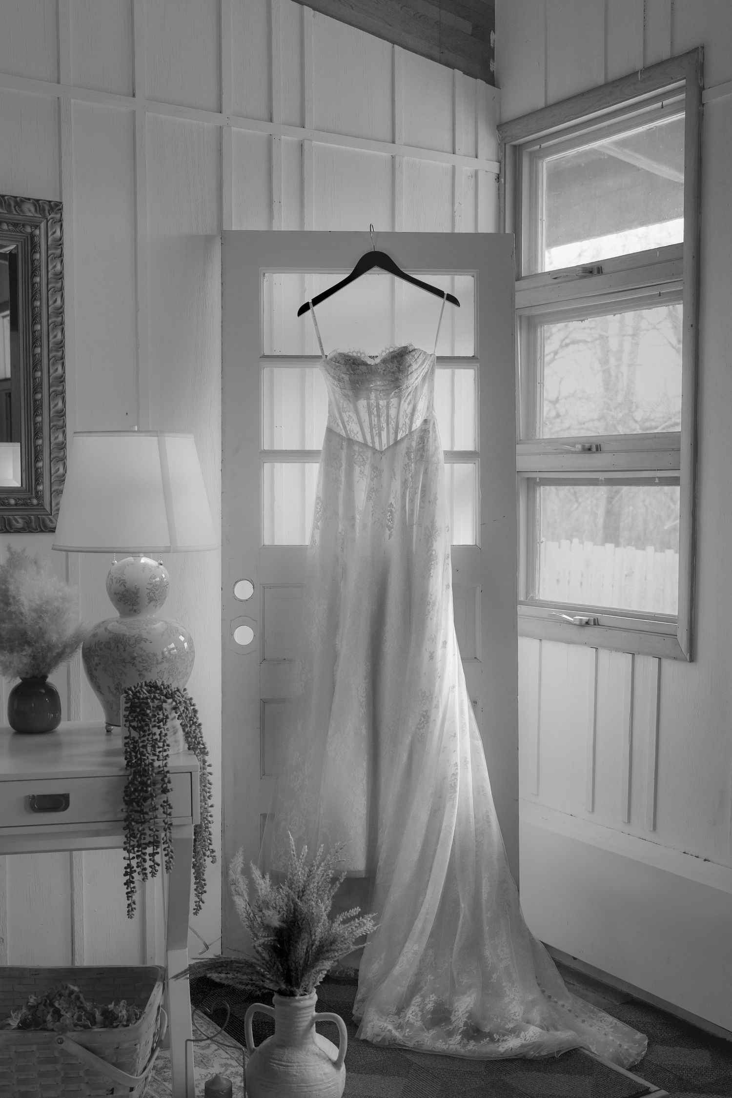 Black and white photo of an elegant lace wedding dress hanging on a door in a bright rustic room with windows and decor.