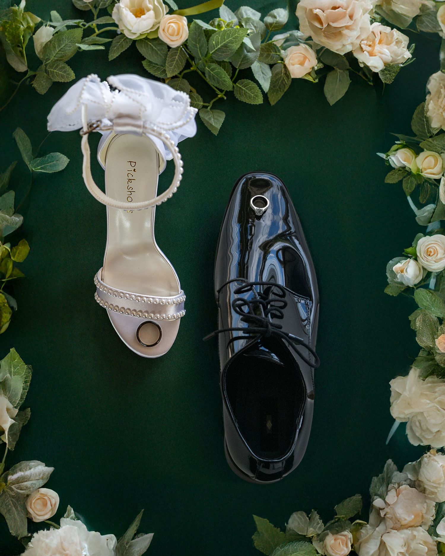 Wedding shoes flat lay featuring a white bridal heel and black groom dress shoe surrounded by cream roses on green.