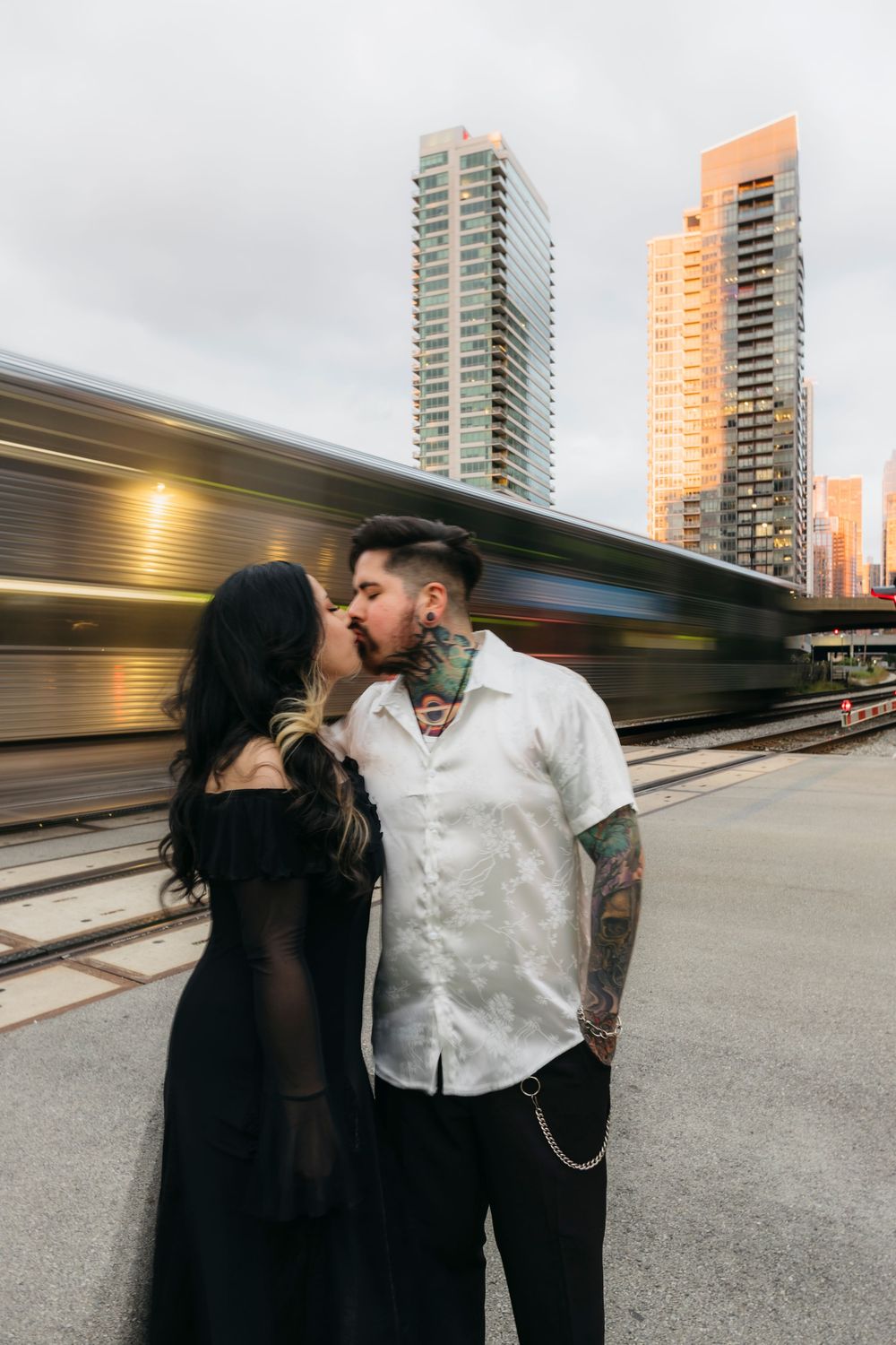 A tattooed couple shares a kiss on a train platform with blurred passing train and Chicago city skyscrapers in background at Fulton Market
