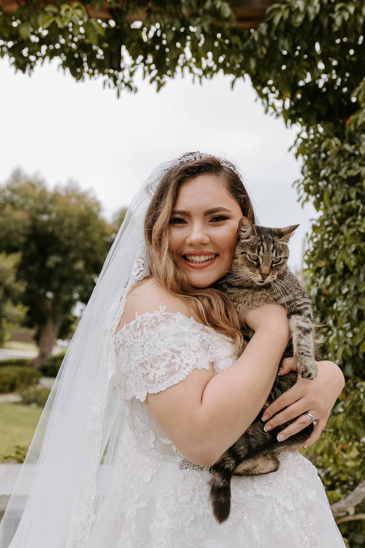 Smiling bride in lace off-shoulder gown and veil holds tabby cat outdoors under floral arbor at The Thursday Club.