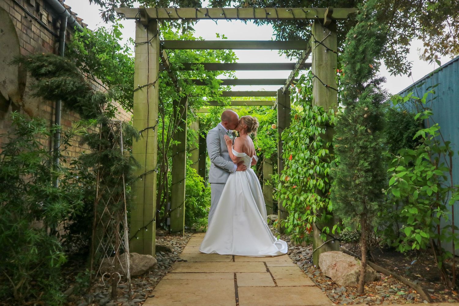 Bride and groom share a romantic kiss under a wooden pergola surrounded by lush green garden foliage on their wedding day.